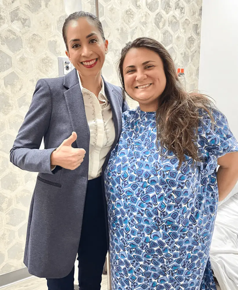 Two smiling women standing close together, one giving a thumbs-up, against a light patterned wall.
