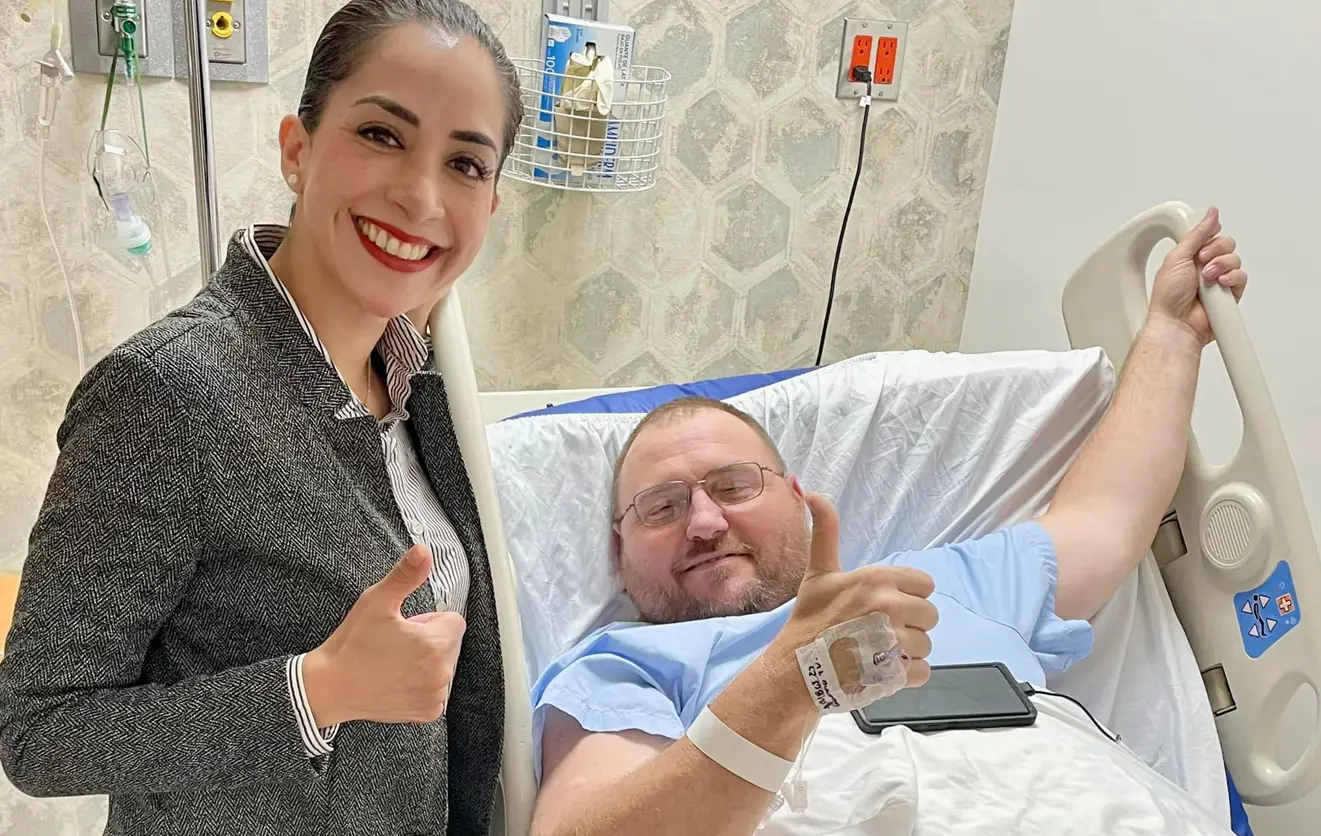 Smiling woman giving thumbs up standing next to a hospital patient in bed who is also giving thumbs up.