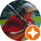 Man wearing a beige cap and red shirt swinging a golf club on a green golf course.