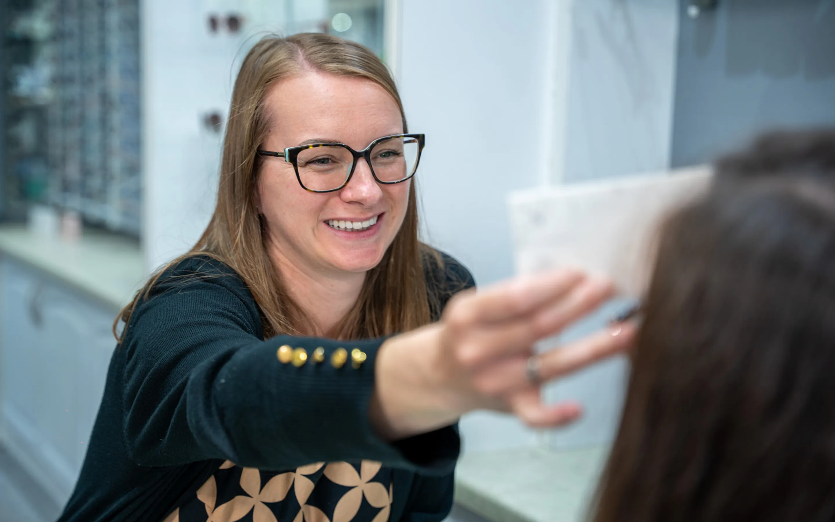 Smiling woman wearing glasses holding an eye test card in front of another person in an optometrist office.