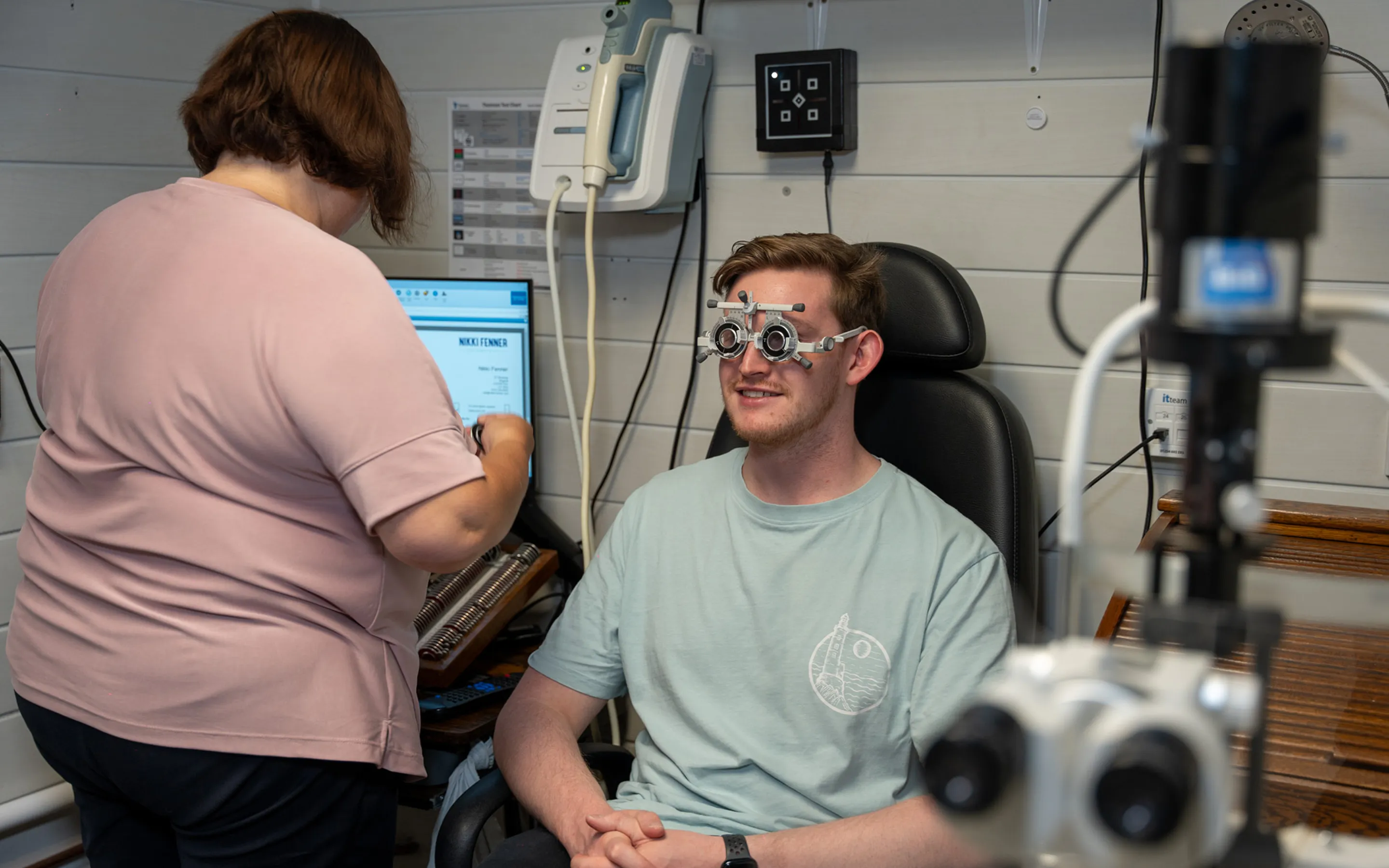 Man sitting in an eye exam chair wearing a phoropter while a technician adjusts lenses nearby.