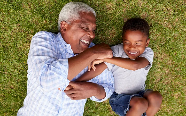 Smiling older man playfully interacting with laughing young boy while lying on green grass.