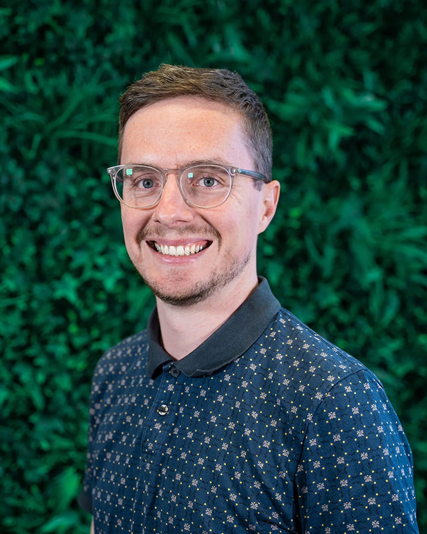 Smiling man with short brown hair and glasses wearing a patterned blue polo shirt in front of a green leafy background.