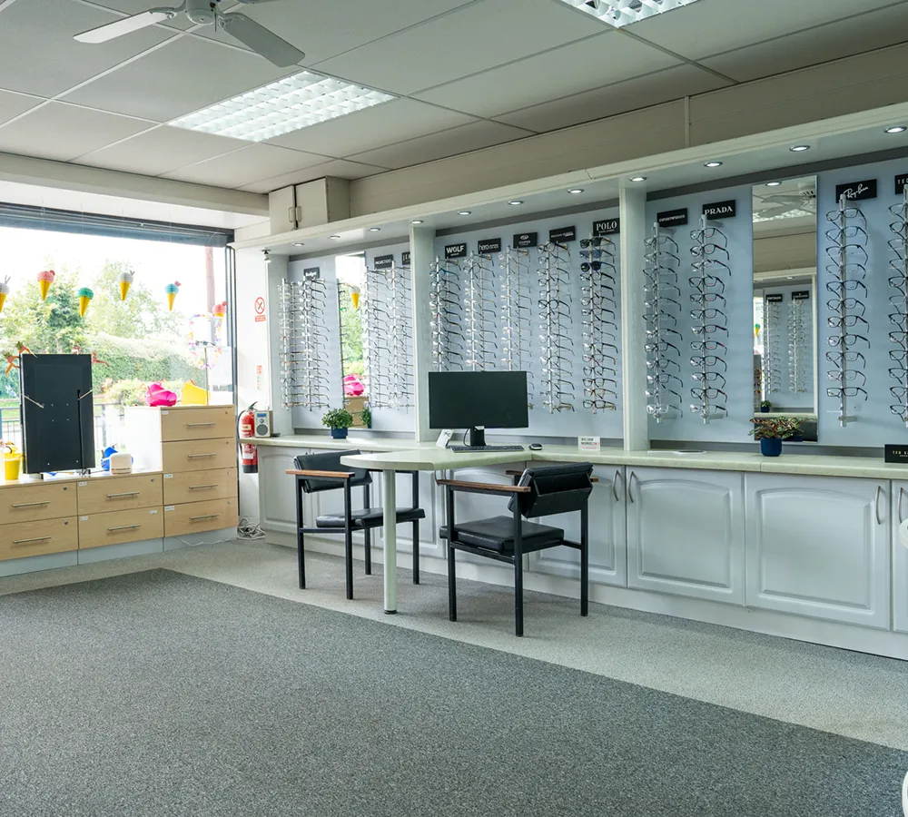 Bright optician’s office with multiple eyeglass frames on wall displays, two chairs, a desk with a computer monitor, and large windows.