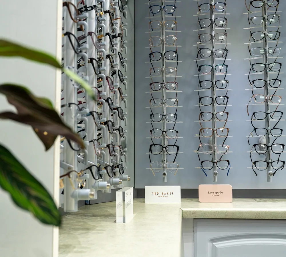 Display of various eyeglasses frames on racks with Ted Baker and Kate Spade brand labels on a countertop.
