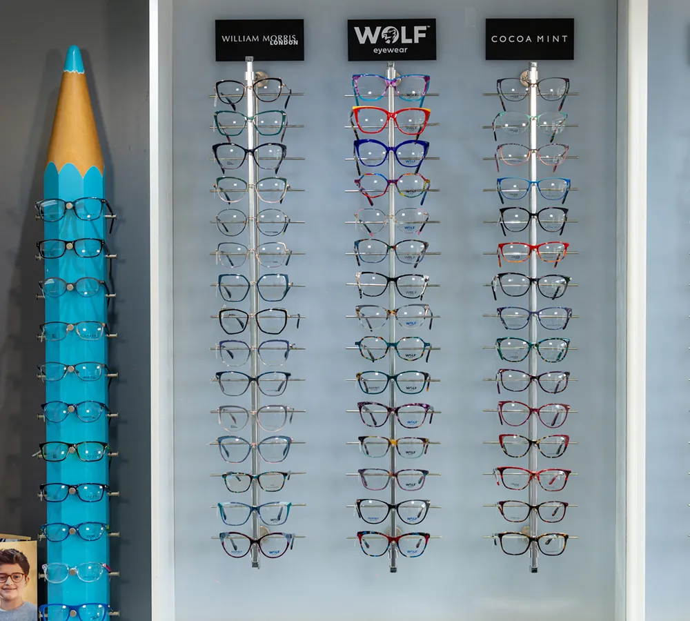 Display of various eyeglasses on racks labeled William Morris, Wolf eyewear, and Cocoa Mint against a light gray background.