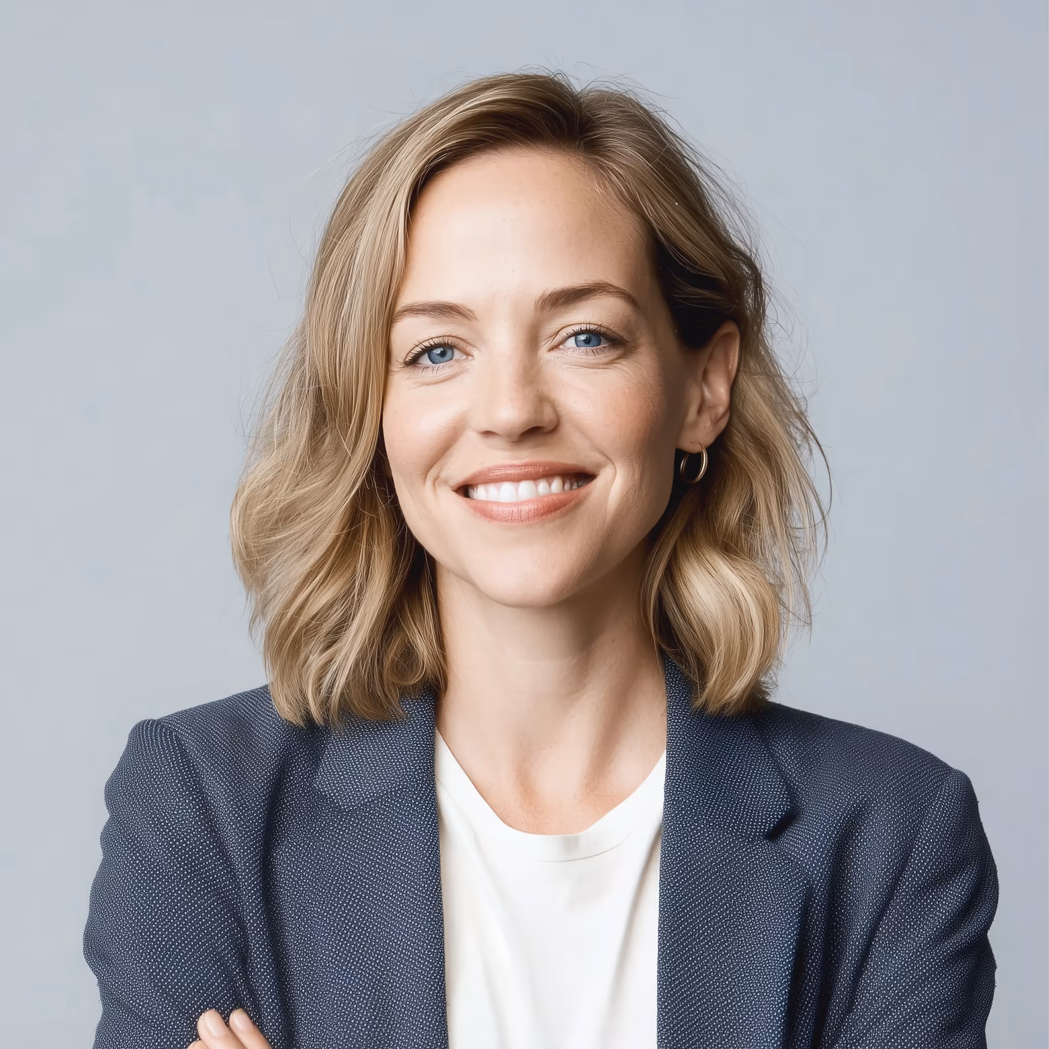 Smiling woman with blonde hair wearing a navy blazer and white shirt against a gray background.