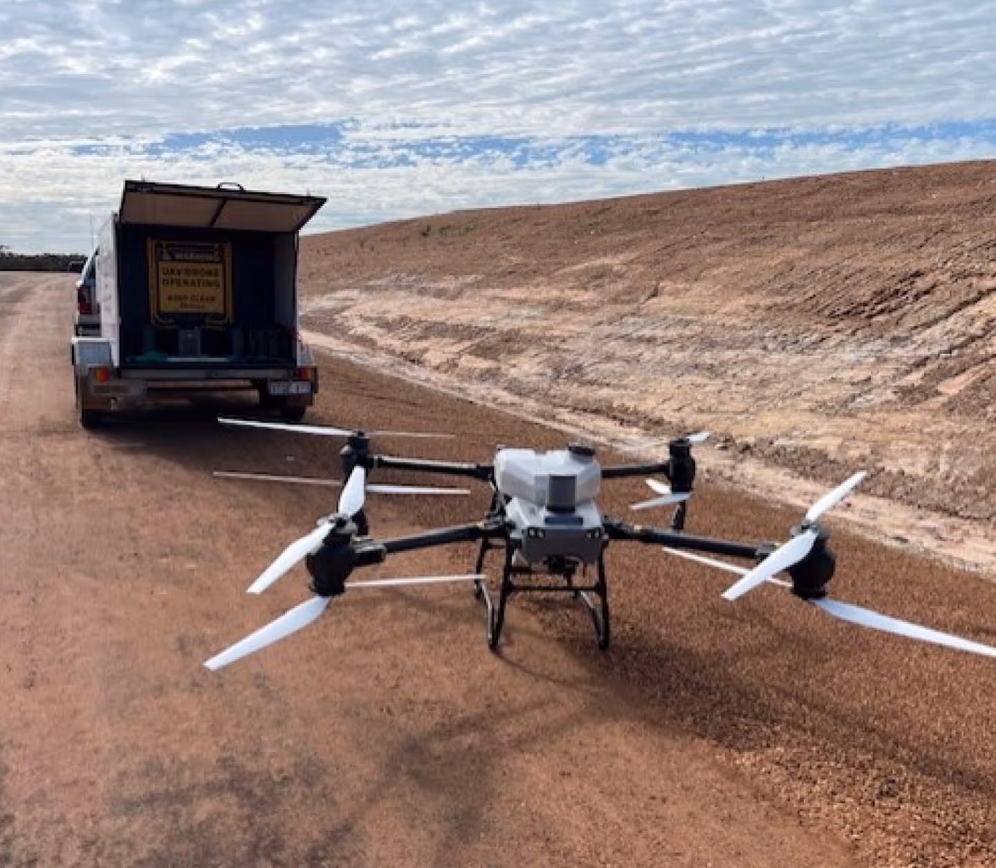 Large quadcopter drone on a dirt road near an open trailer with operational warning sign, against a partly cloudy sky.