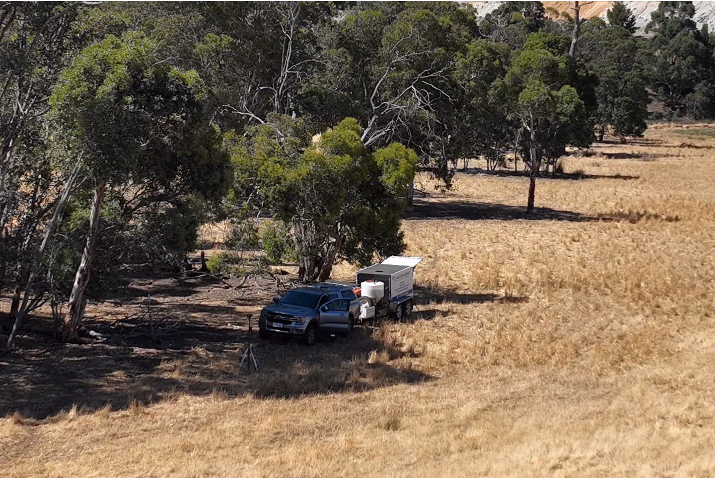 Pickup truck with an open door attached to a small trailer parked at the edge of a forested area next to a dry grassy field.