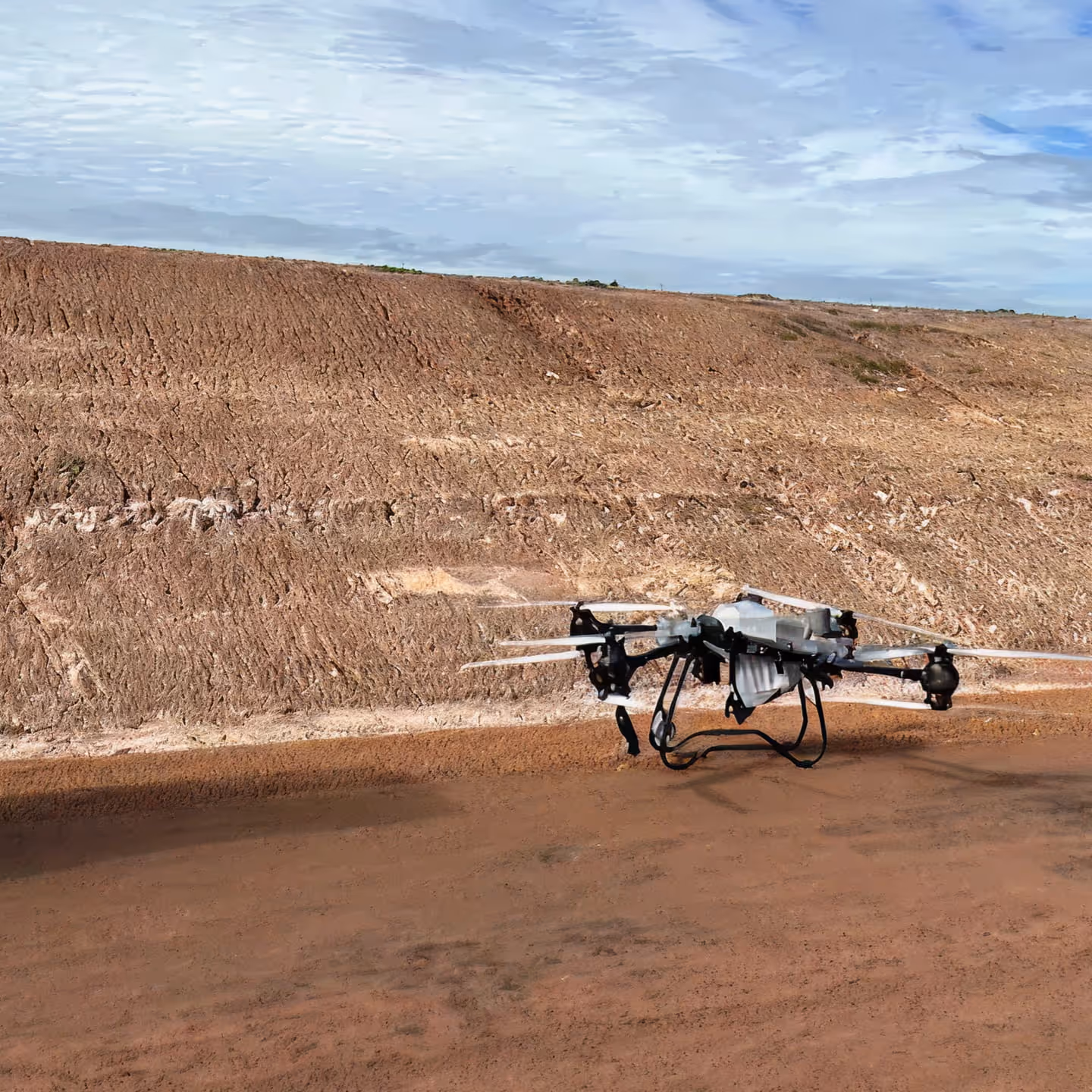 Black and silver drone hovering above dry, sandy ground with a dirt embankment under a partly cloudy sky.