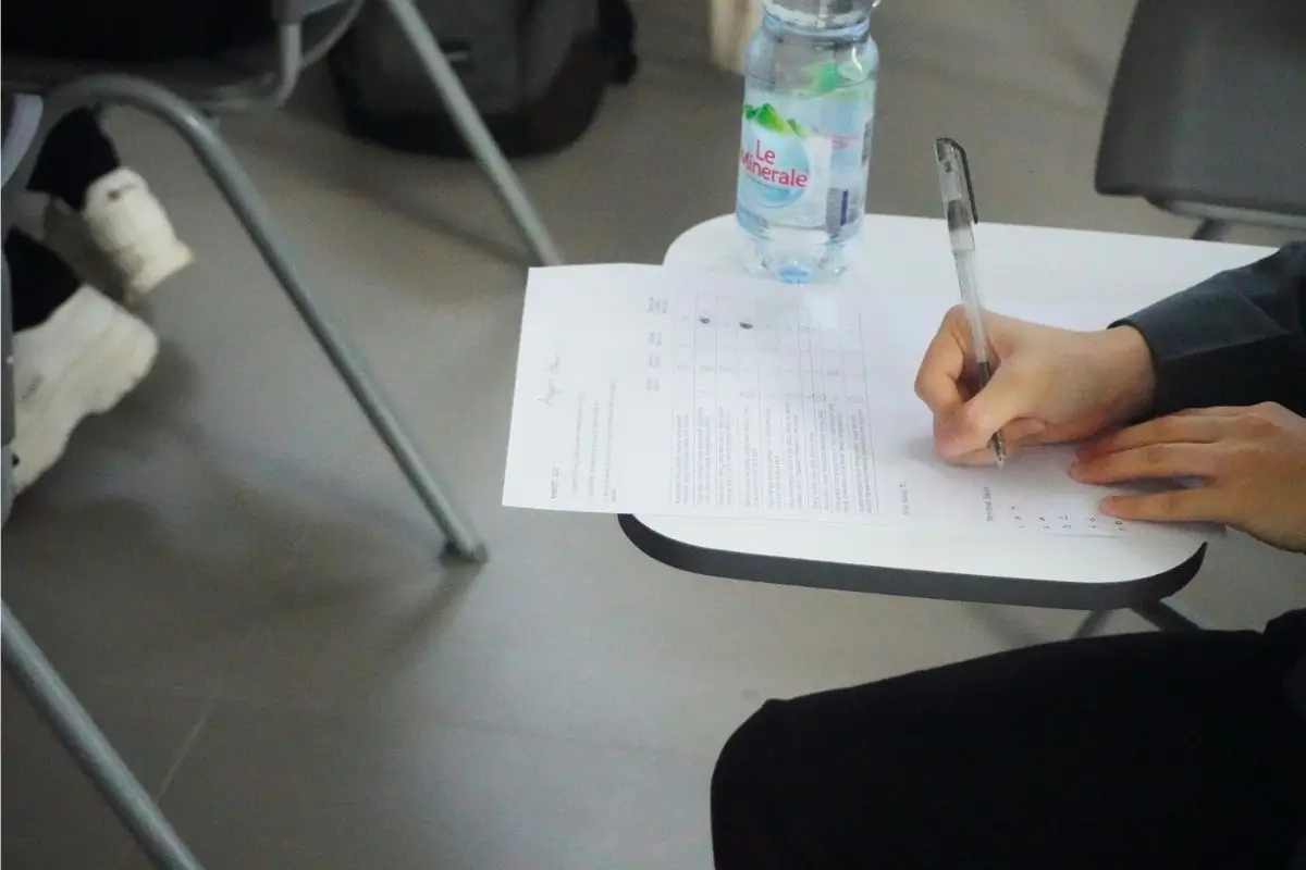 An anxious student sits at a table, focused on their Economics exam, with a pen in hand and paper in front of them.