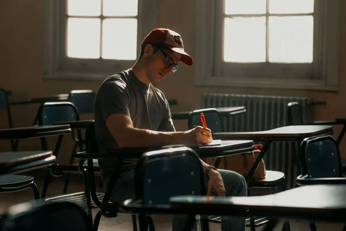 A male student in a cap writes notes on paper while studying alone in a university classroom.