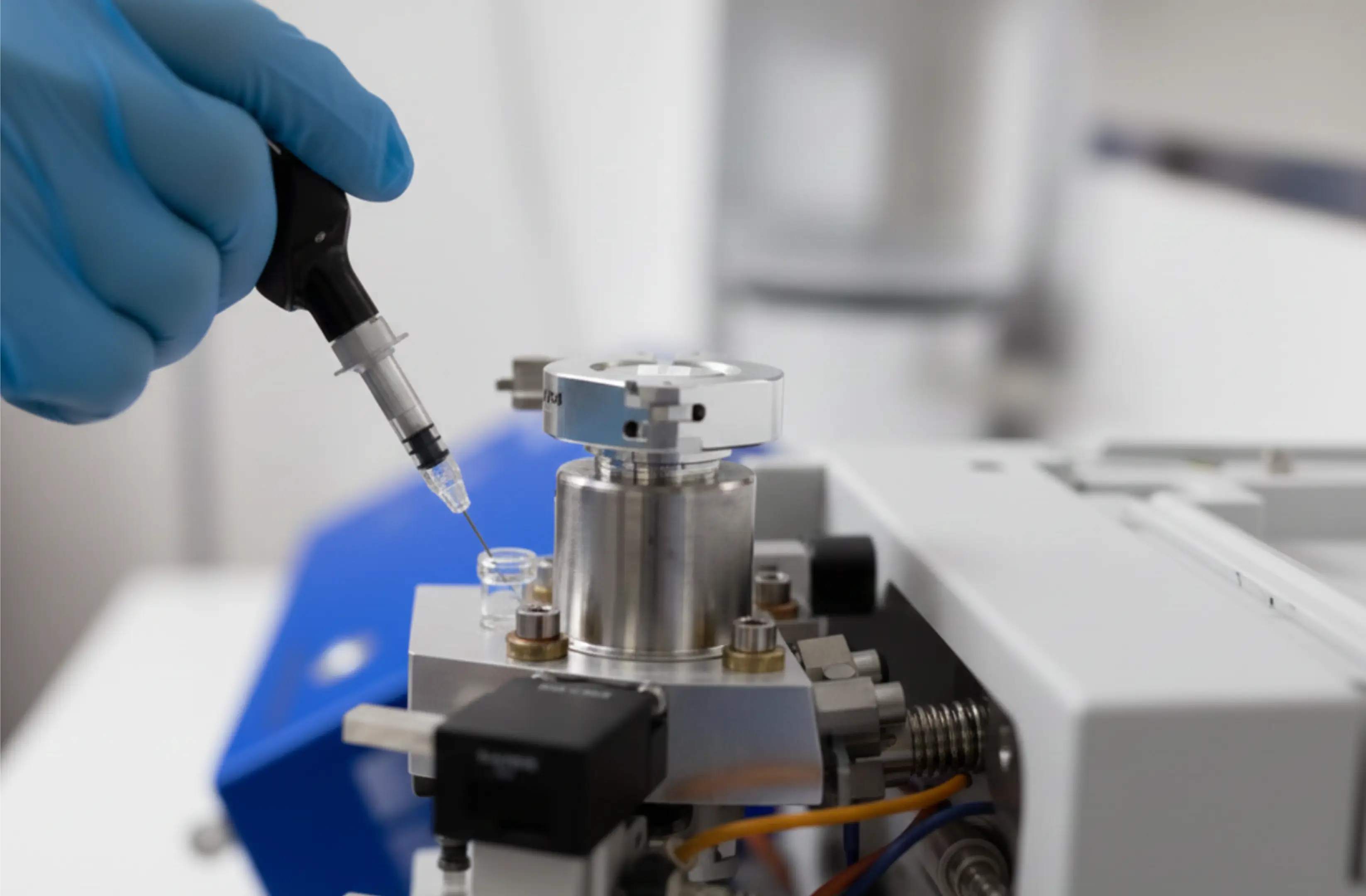 Gloved hand holding a syringe near a metal component of laboratory machinery.