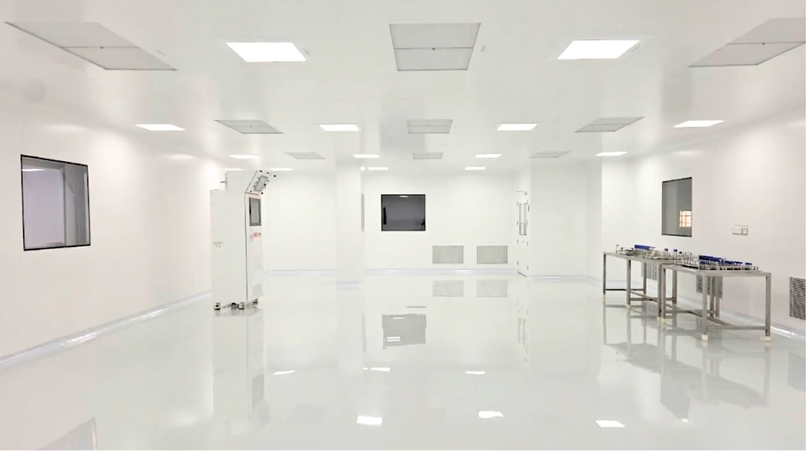 Bright, clean laboratory room with white walls, reflective floor, ceiling lights, a machine on the left, and metal tables with lab equipment on the right.