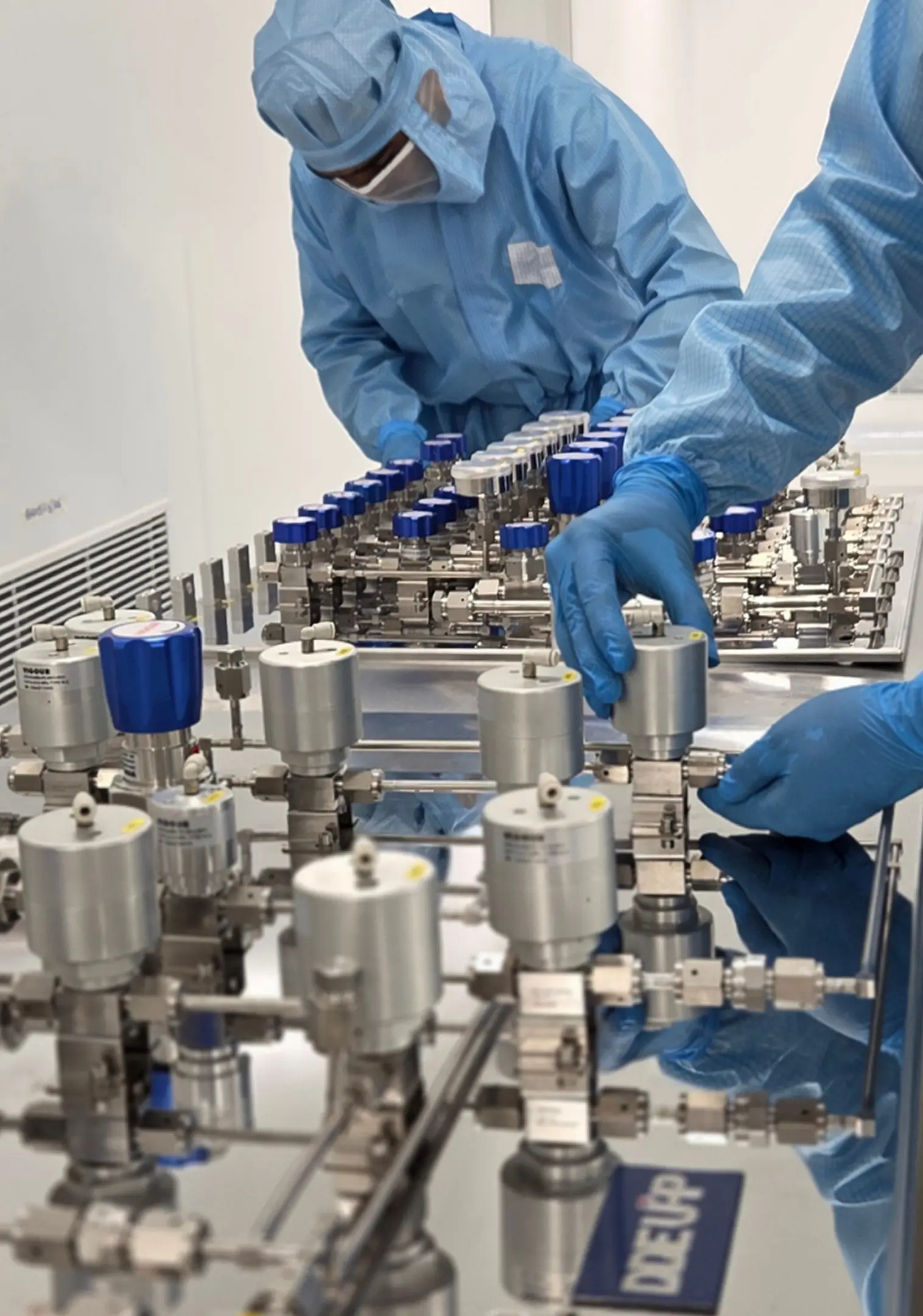 Technicians in blue protective suits and gloves working on a complex array of stainless steel valves and fittings in a laboratory or cleanroom.