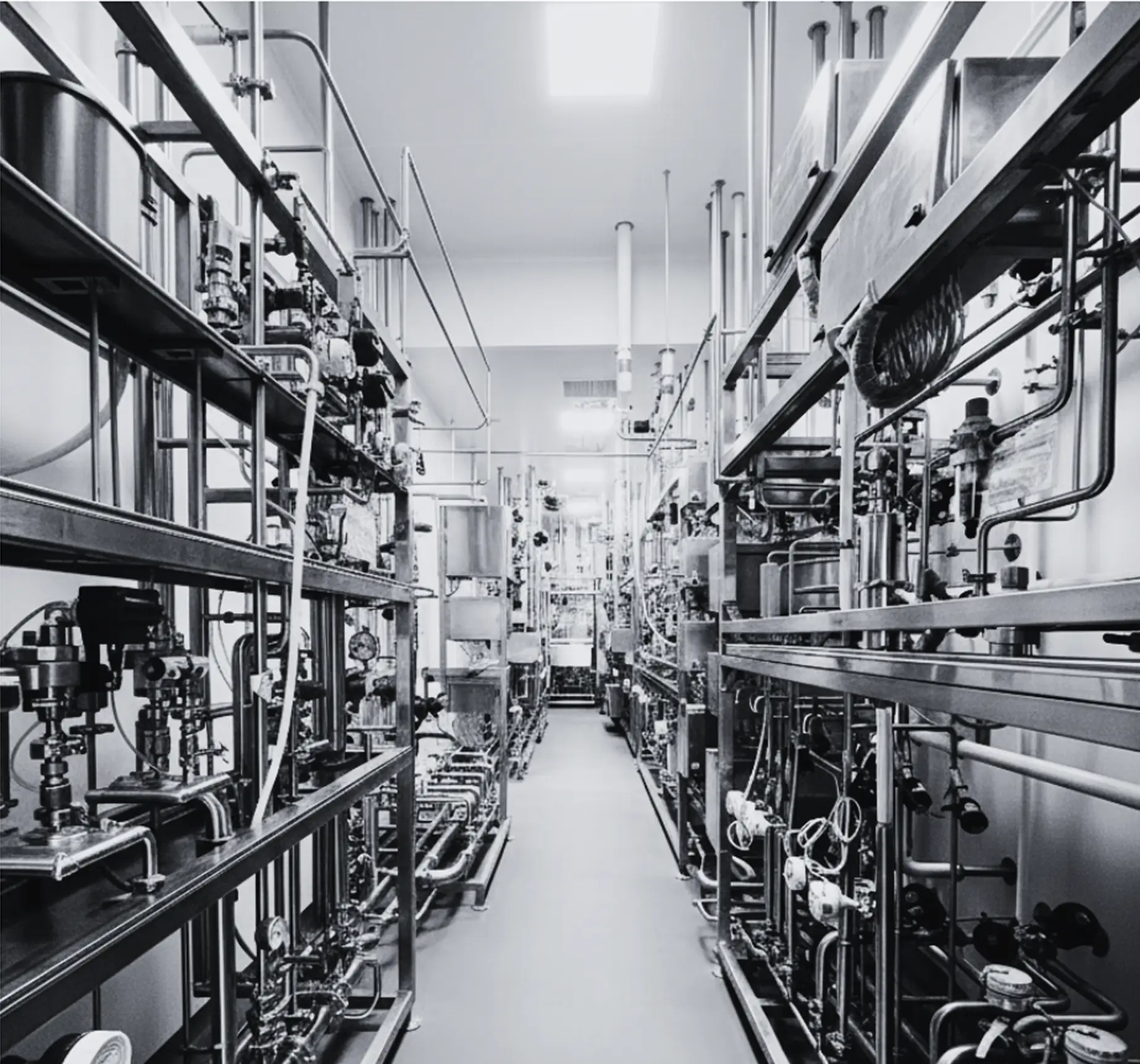 Narrow aisle in a laboratory or industrial facility with stainless steel pipes, gauges, and machinery on both sides.