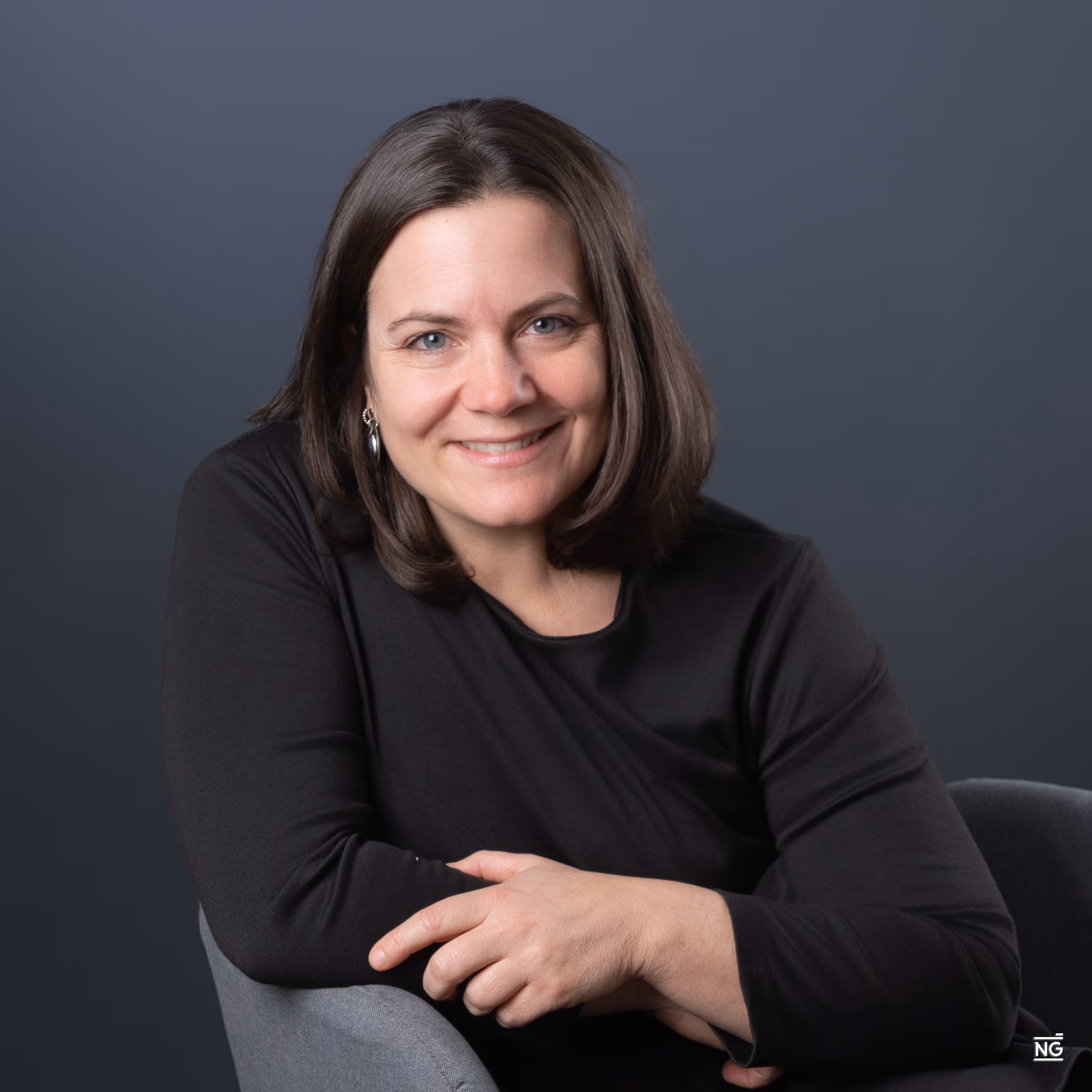 Smiling woman with shoulder-length brown hair wearing a black top, sitting on a gray chair against a dark background.