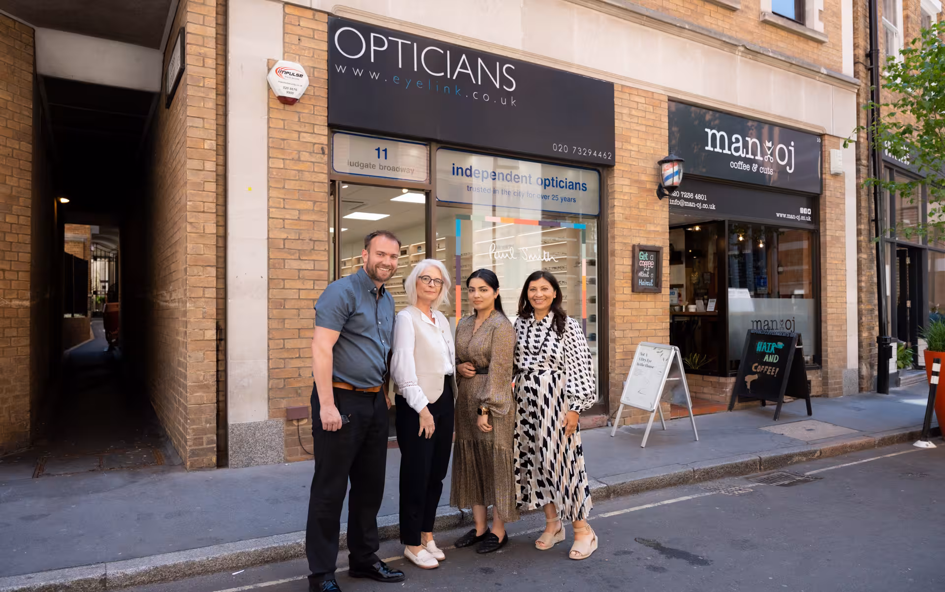 Four people standing and smiling outside an opticians shop on a city street.