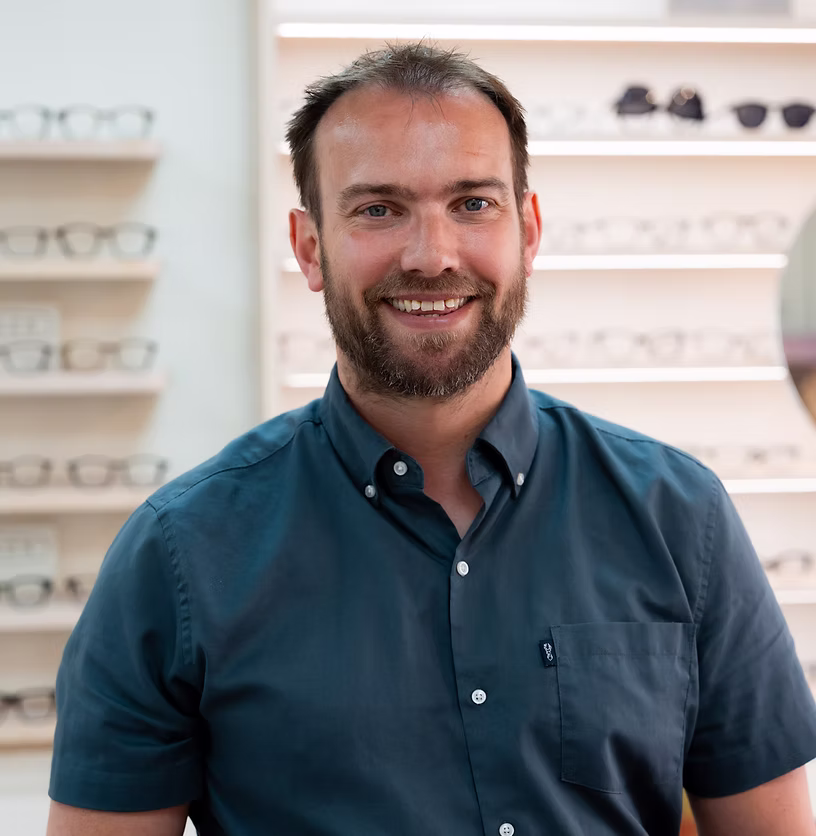 Smiling man with beard in a dark blue shirt stands in front of shelves displaying eyeglasses.