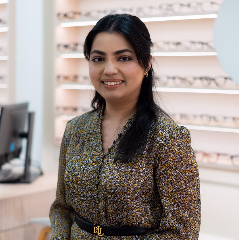 Smiling woman with long dark hair wearing a floral top, standing in an eyewear store.