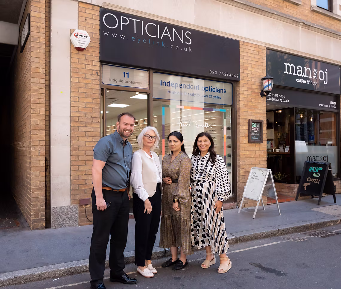 Four people standing outside an optician shop on a city sidewalk on a sunny day.