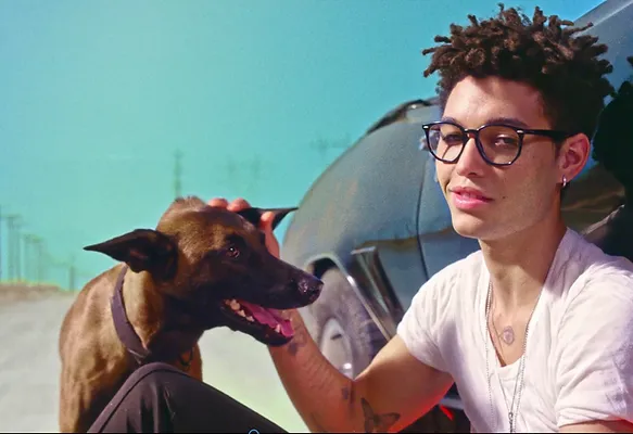 Young man with glasses sitting by a car, petting a brown dog on a sunny road.