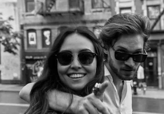 Smiling woman wearing sunglasses points at camera while man with sunglasses hugs her from behind outdoors.