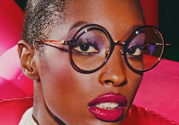 Close-up of a person with round glasses, purple eye shadow, and glossy dark pink lipstick.