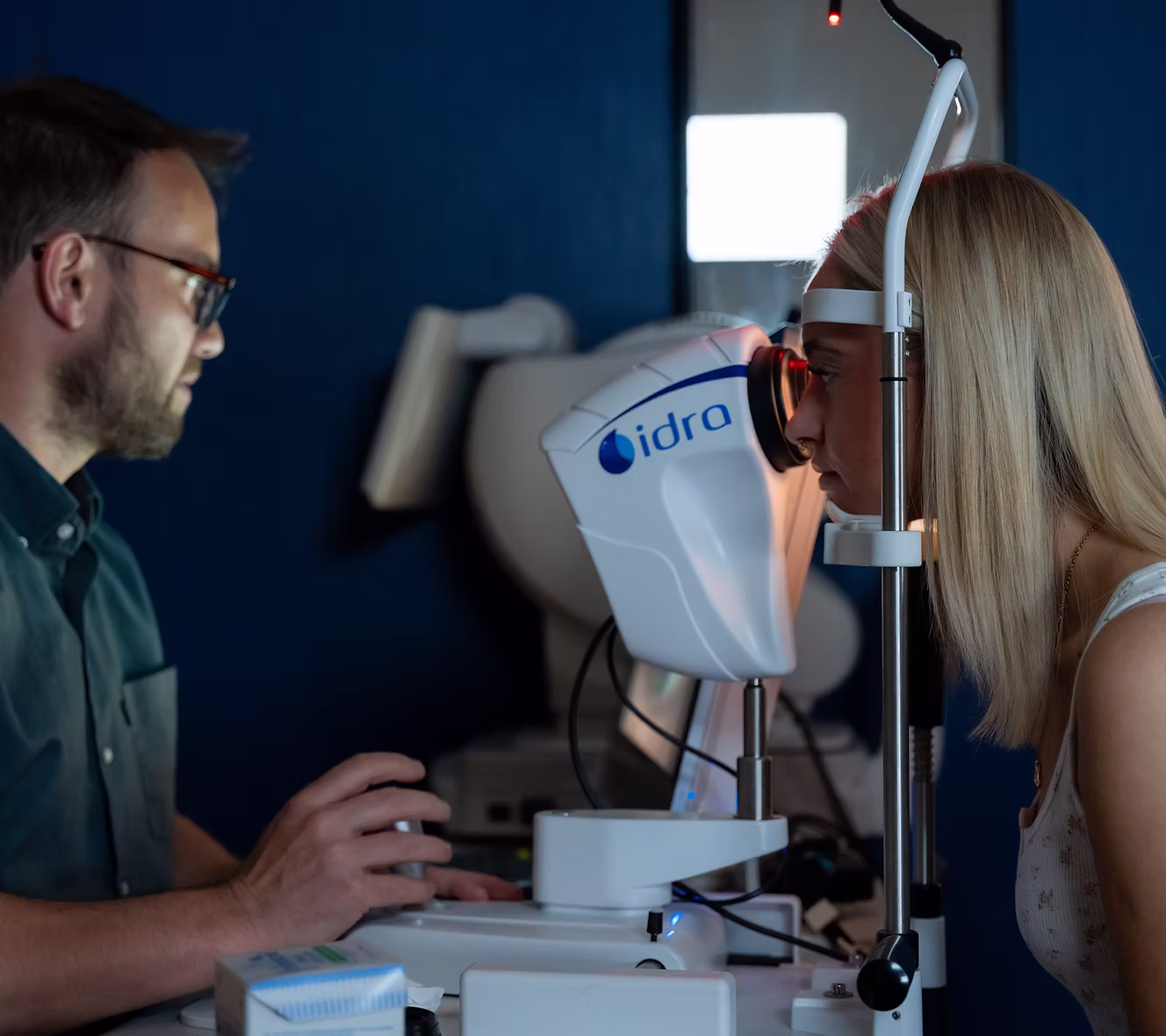 Woman undergoing eye exam using a slit lamp machine operated by an eye care professional.