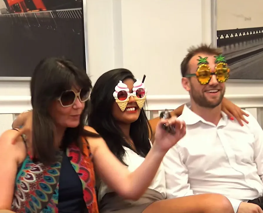 Three adults sitting closely, wearing novelty sunglasses shaped like pineapples and ice cream cones, smiling.
