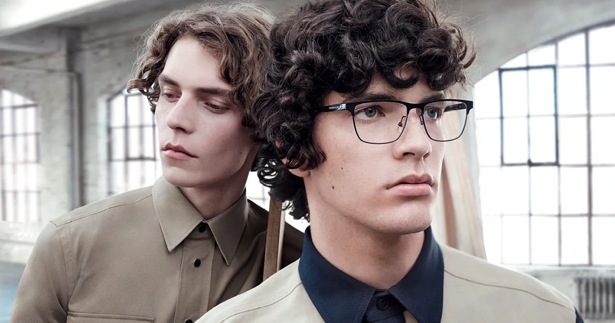 Two young men with curly hair indoors, one wearing glasses and a beige shirt with a dark collar.