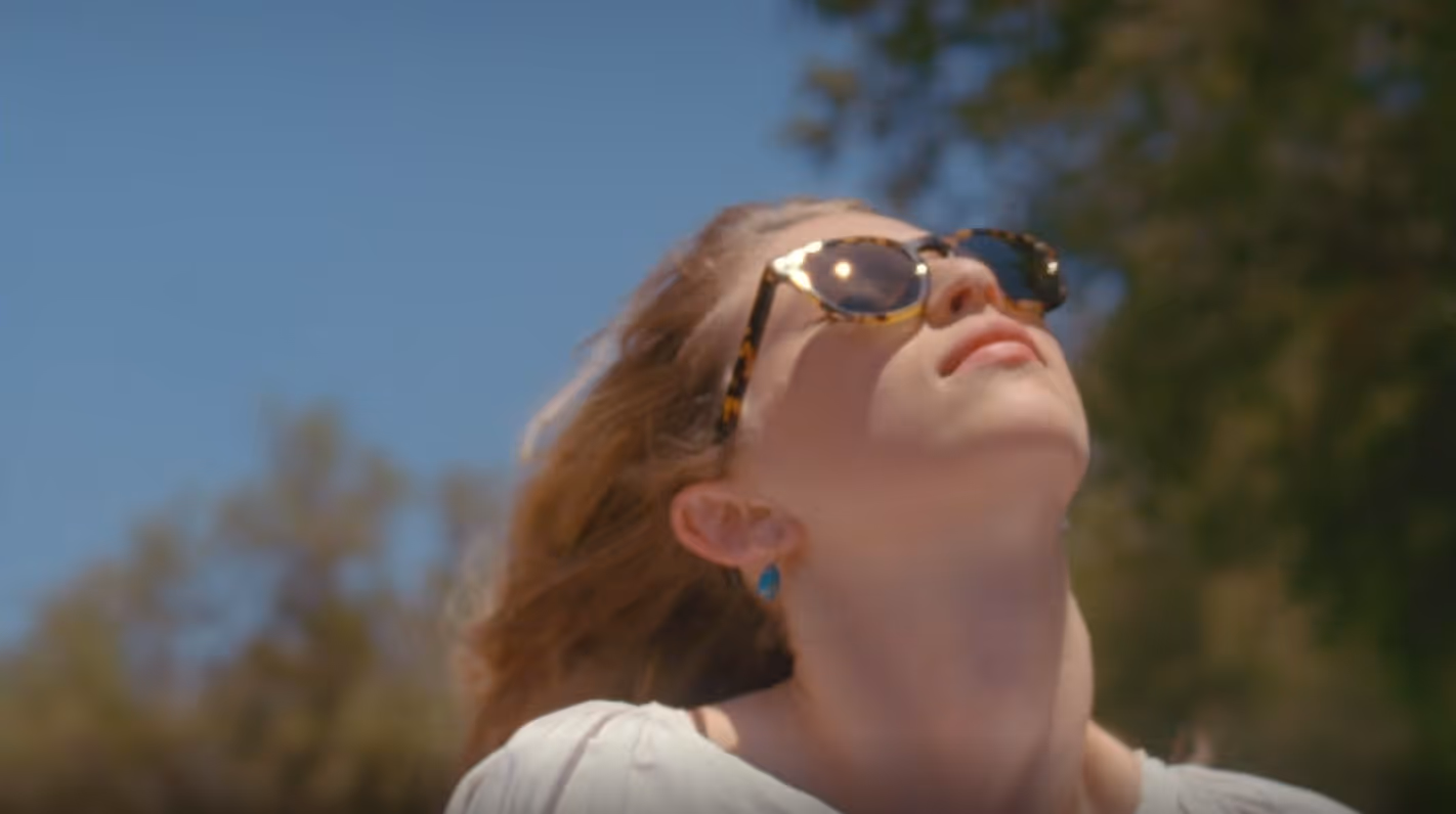 Woman with sunglasses looks up toward the sky with trees blurred in the background.