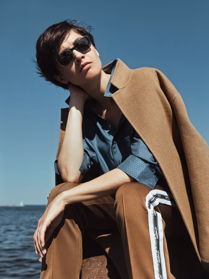 Person wearing sunglasses and a brown coat draped over shoulders, seated by a large body of water.