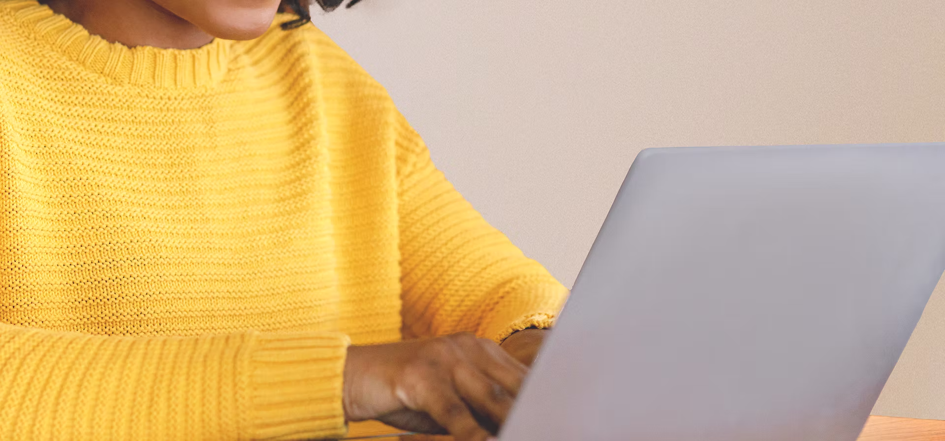 Person wearing a yellow sweater typing on a silver laptop at a wooden table.