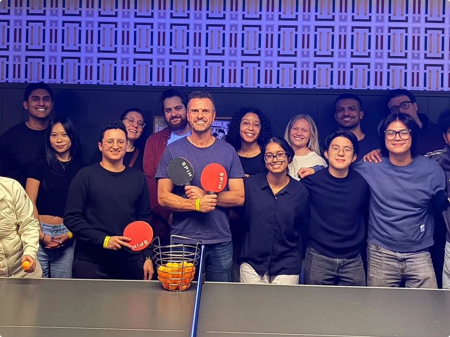 Group of people smiling and posing behind a ping pong table with paddles and ball basket.