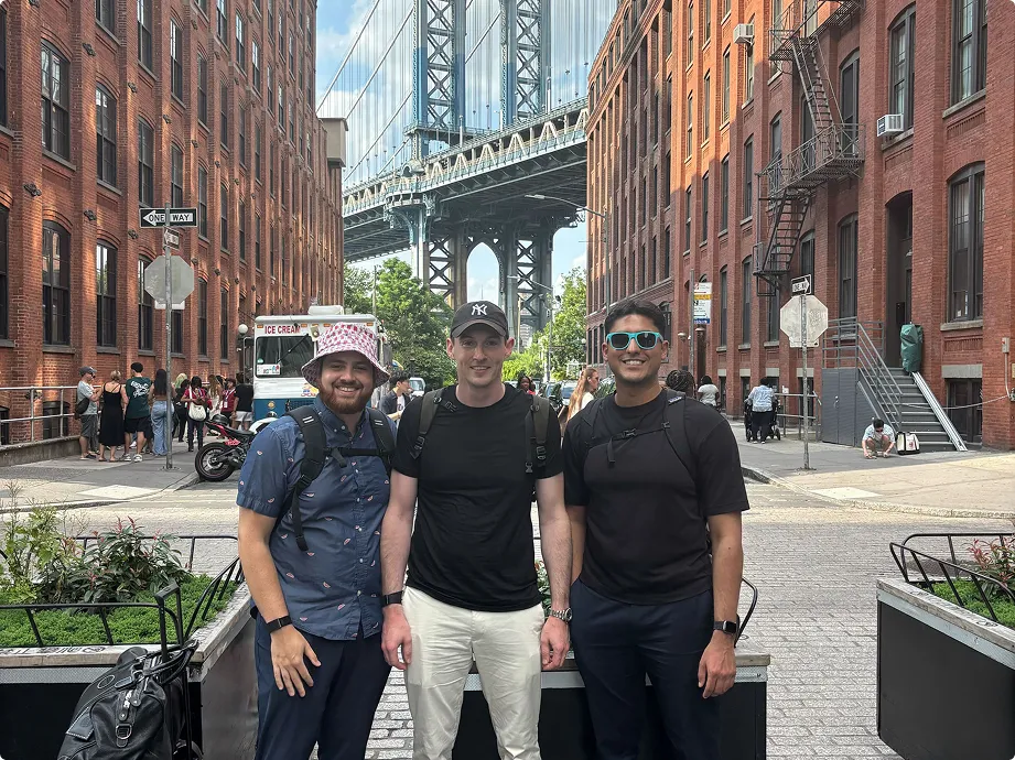 Three men posing in front of Manhattan Bridge between red brick buildings in Dumbo, Brooklyn.