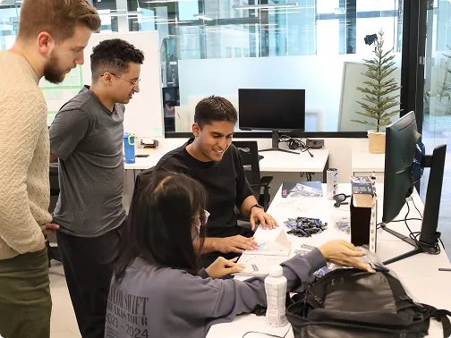 Four people collaborate over documents at a desk with computer monitors in a modern office.