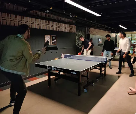 Four men playing ping pong in an indoor recreational room with dark decor and overhead lights.