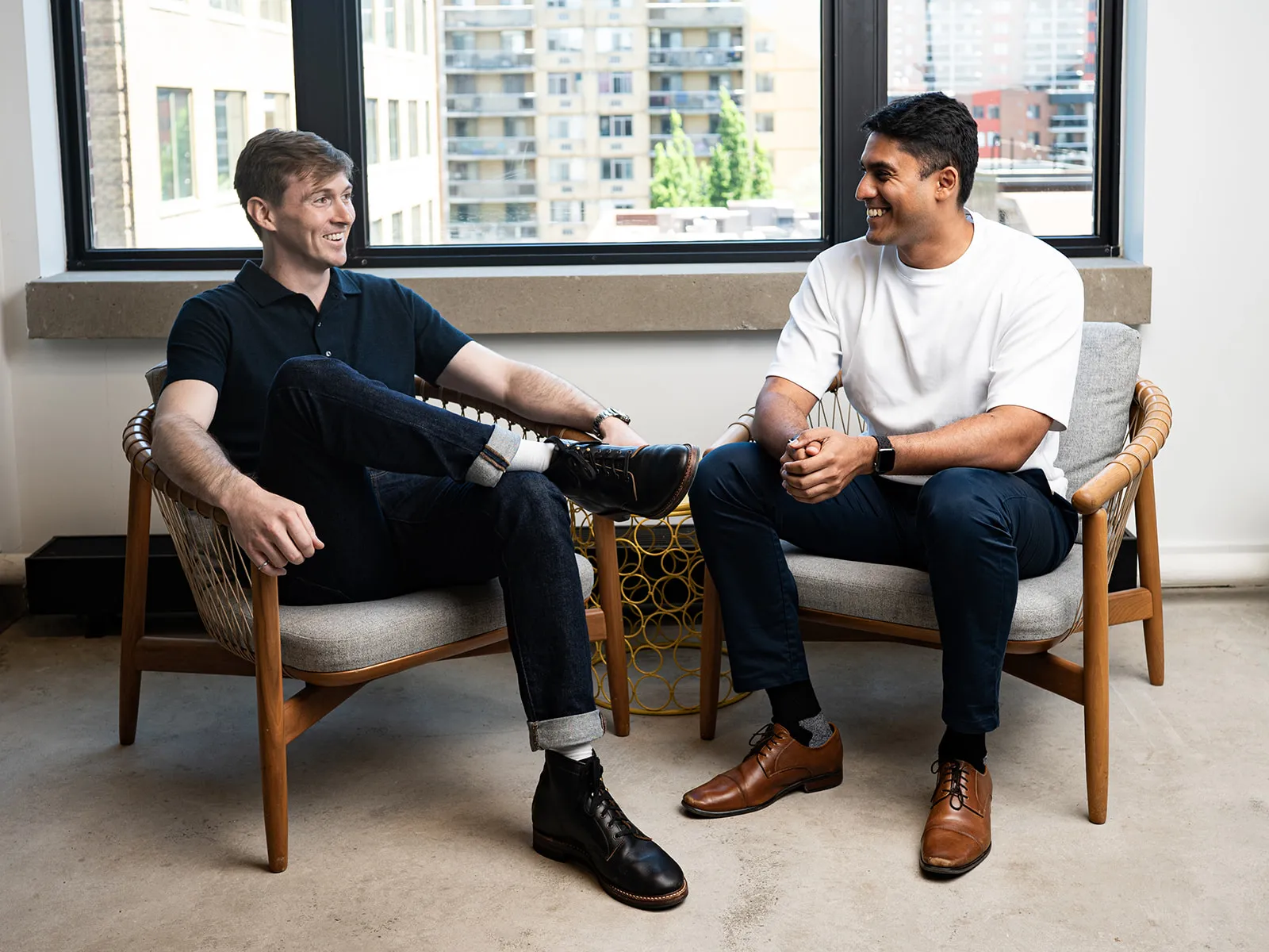Two men sitting in chairs in front of a window, smiling and talking casually indoors.