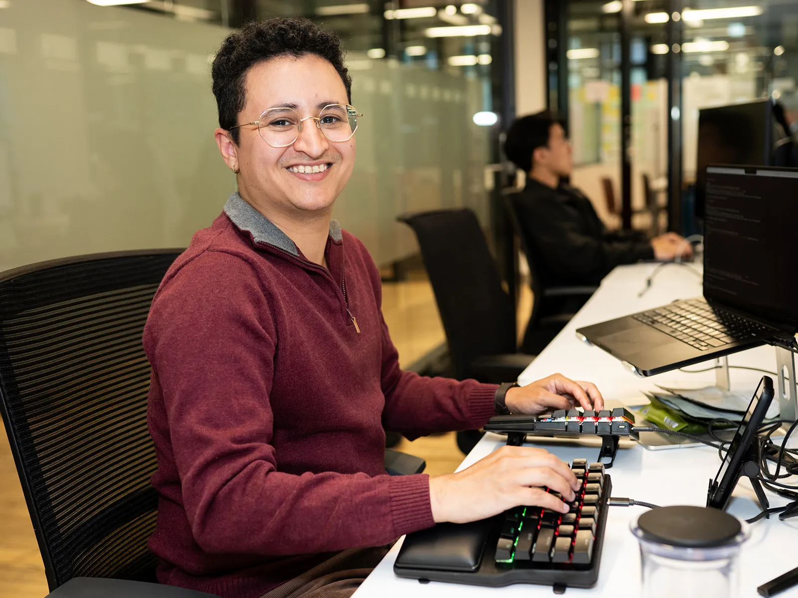 Smiling person wearing glasses and a maroon sweater typing on a split keyboard at a desk.