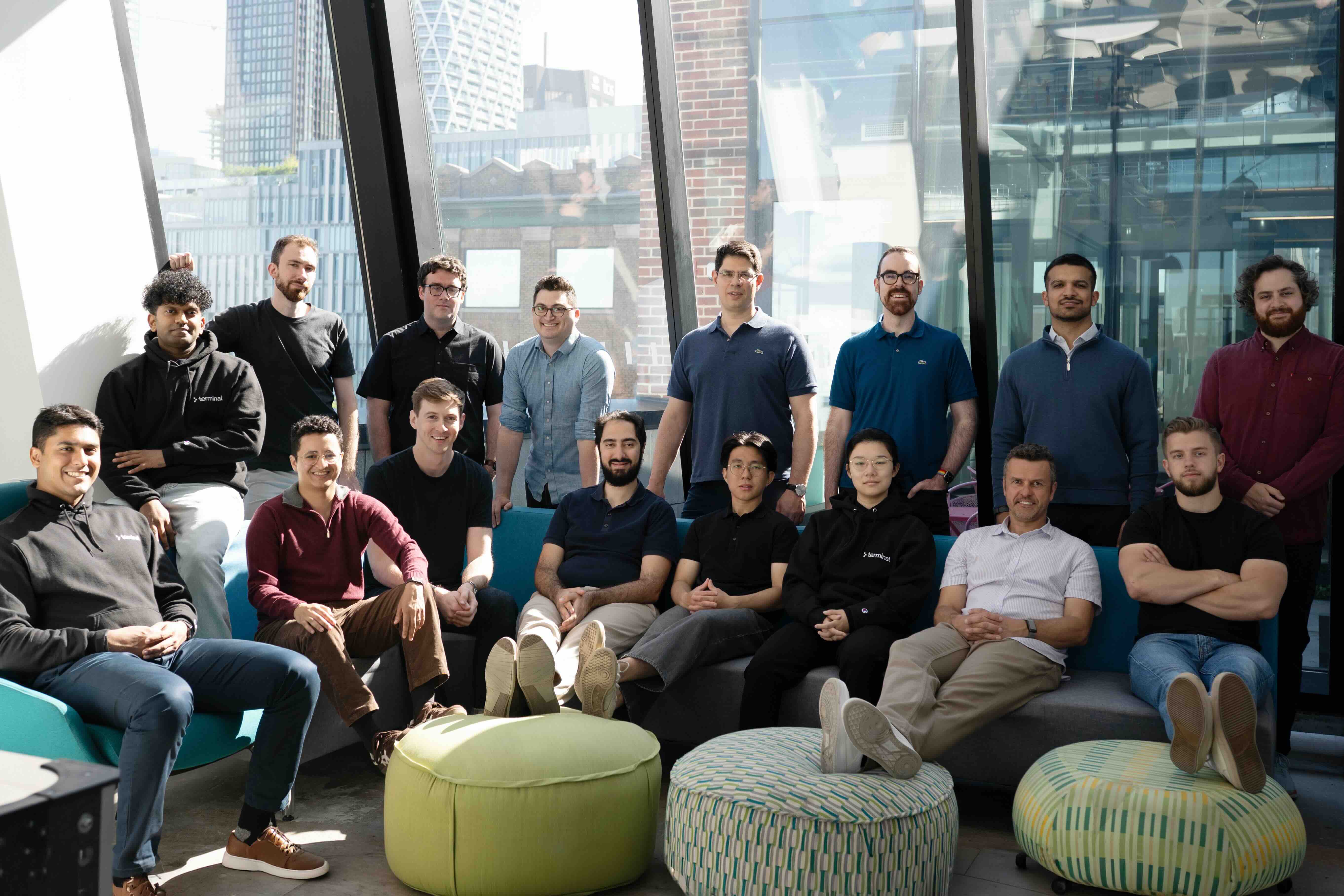 Group of 16 men sitting and standing inside a modern office with large windows showing cityscape.
