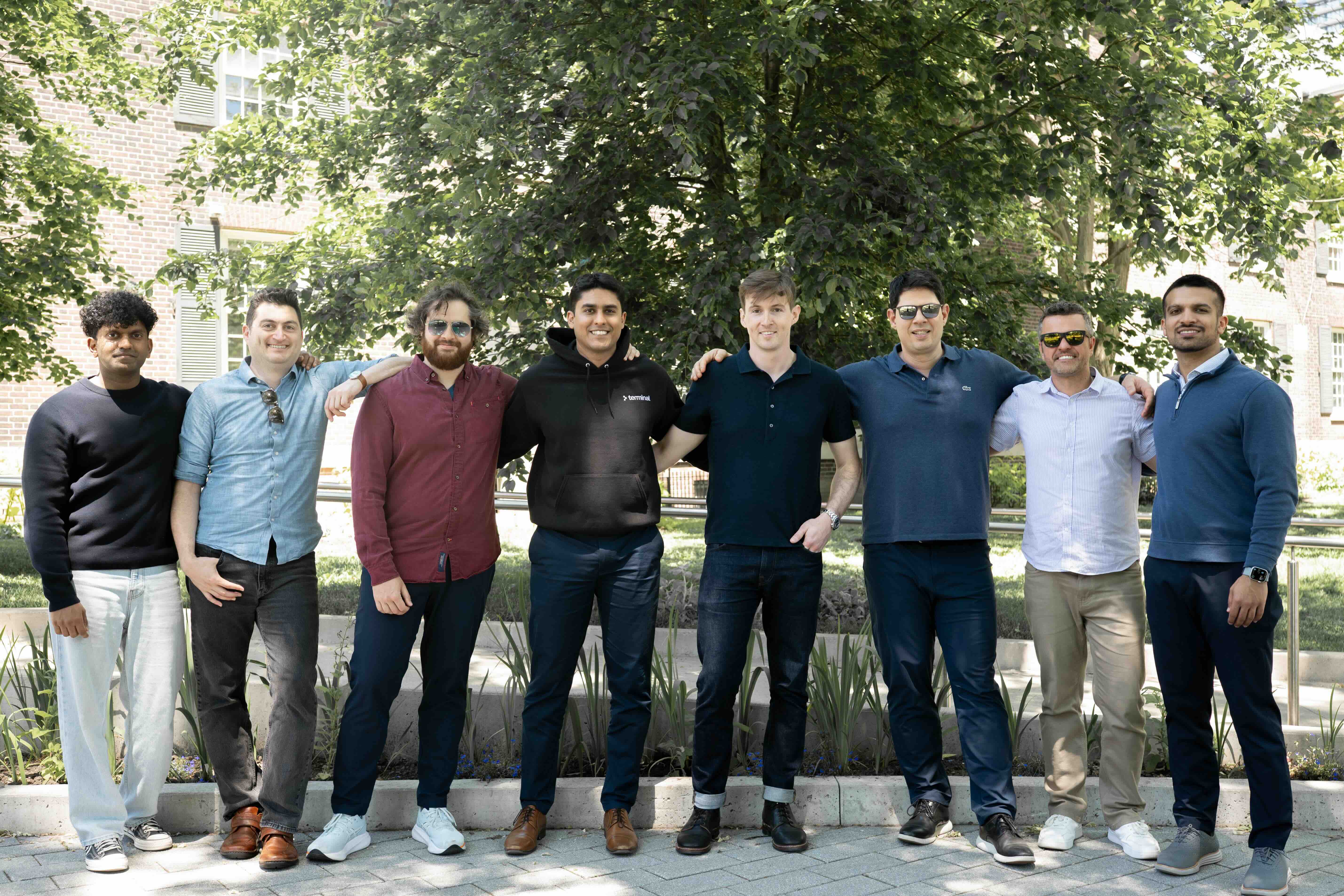 Group of eight men standing outdoors with arms around each other in front of trees and buildings.