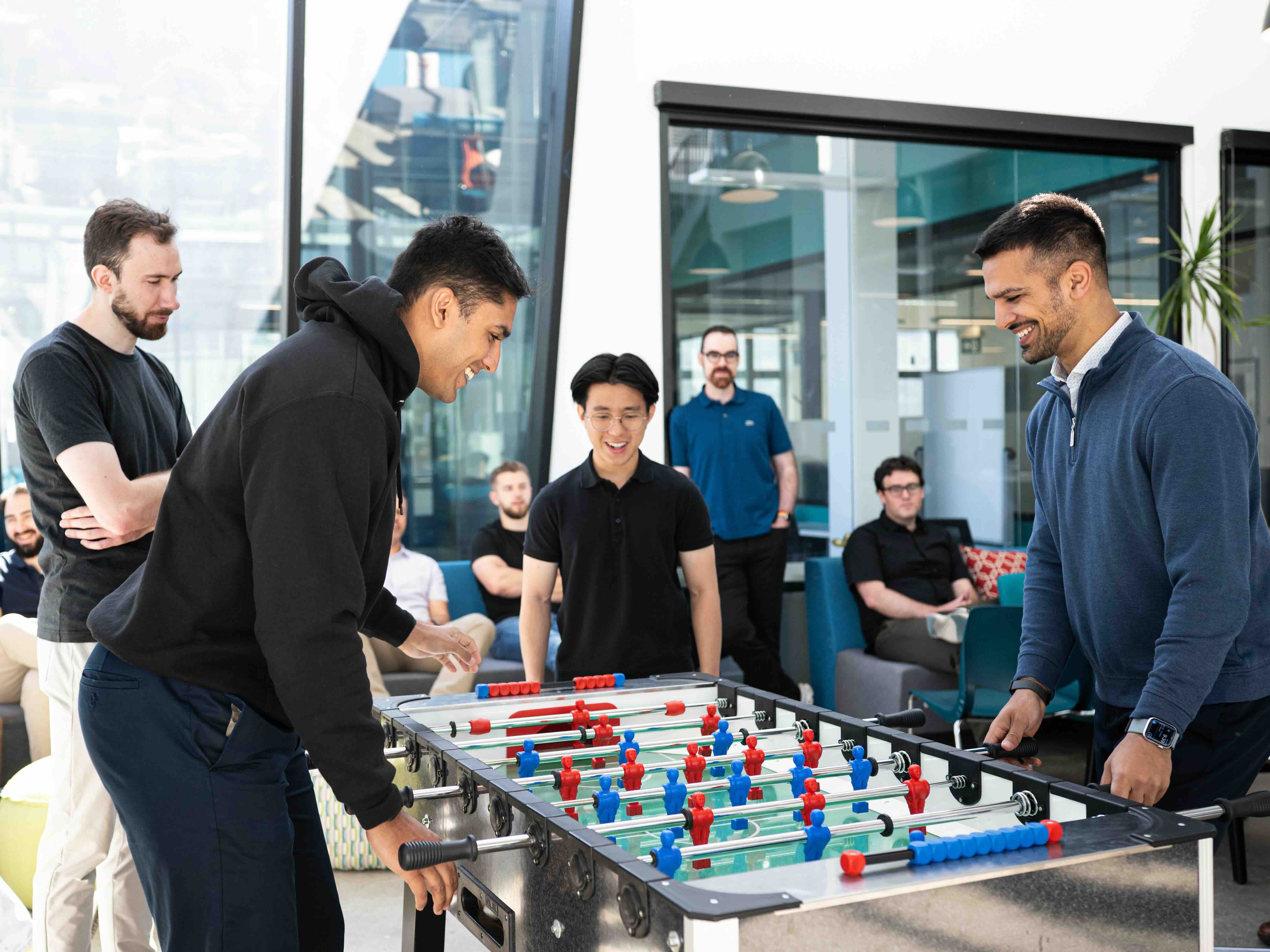 Two men smiling and playing foosball while others watch in a brightly lit modern office.