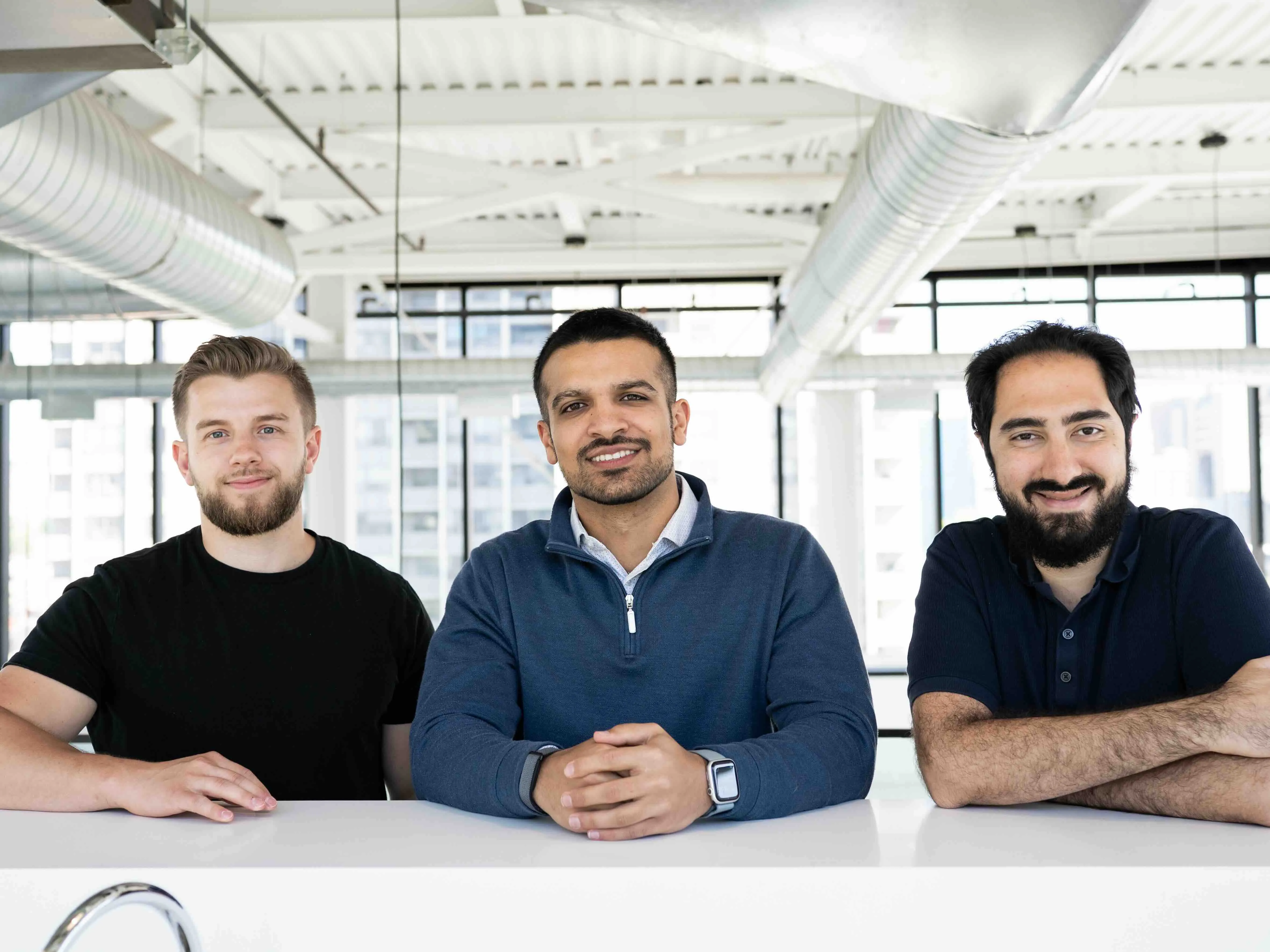 Three men sitting at a white table in a bright modern office, smiling at the camera.