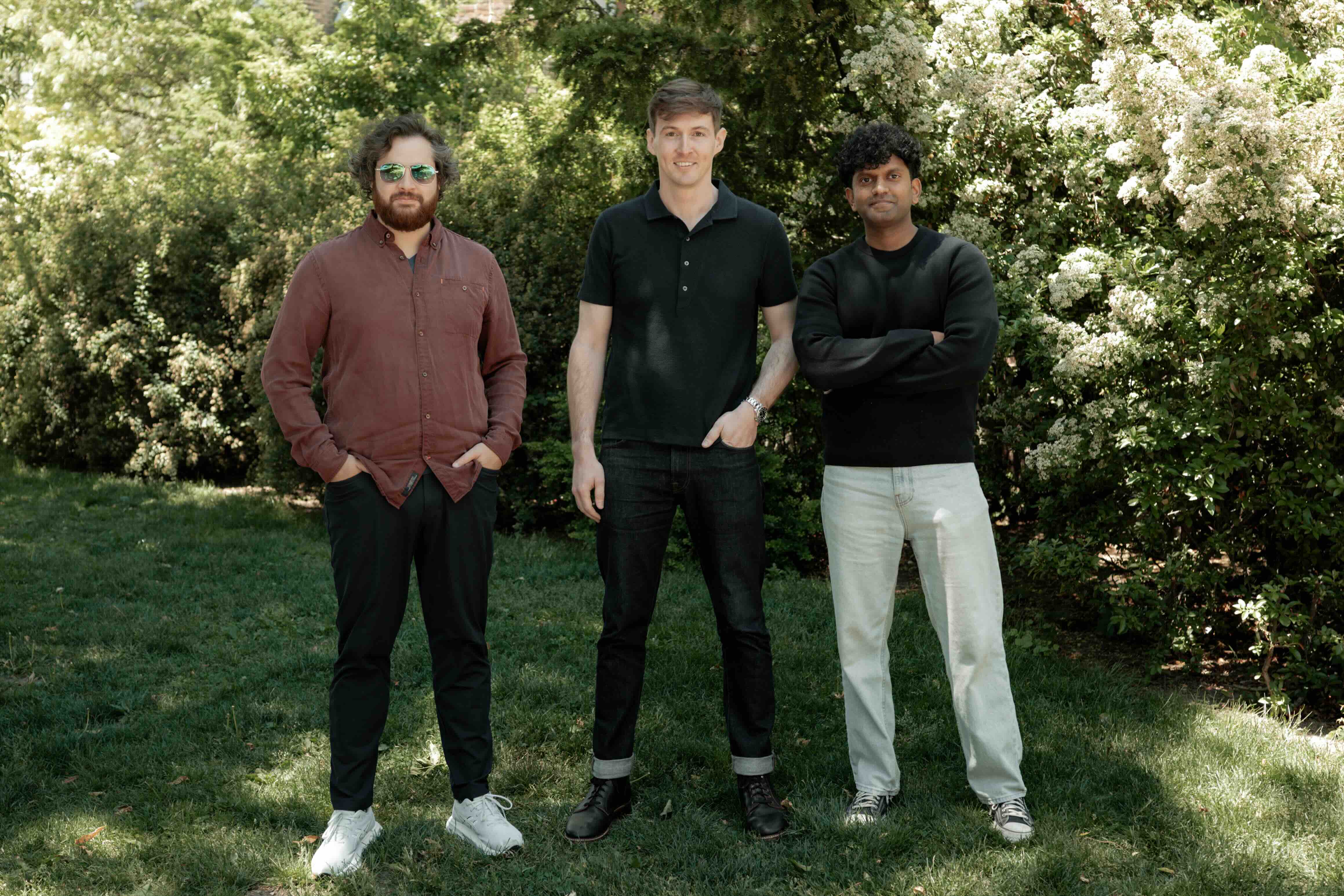 Three men standing outside on grass, with flowering bushes in the background.