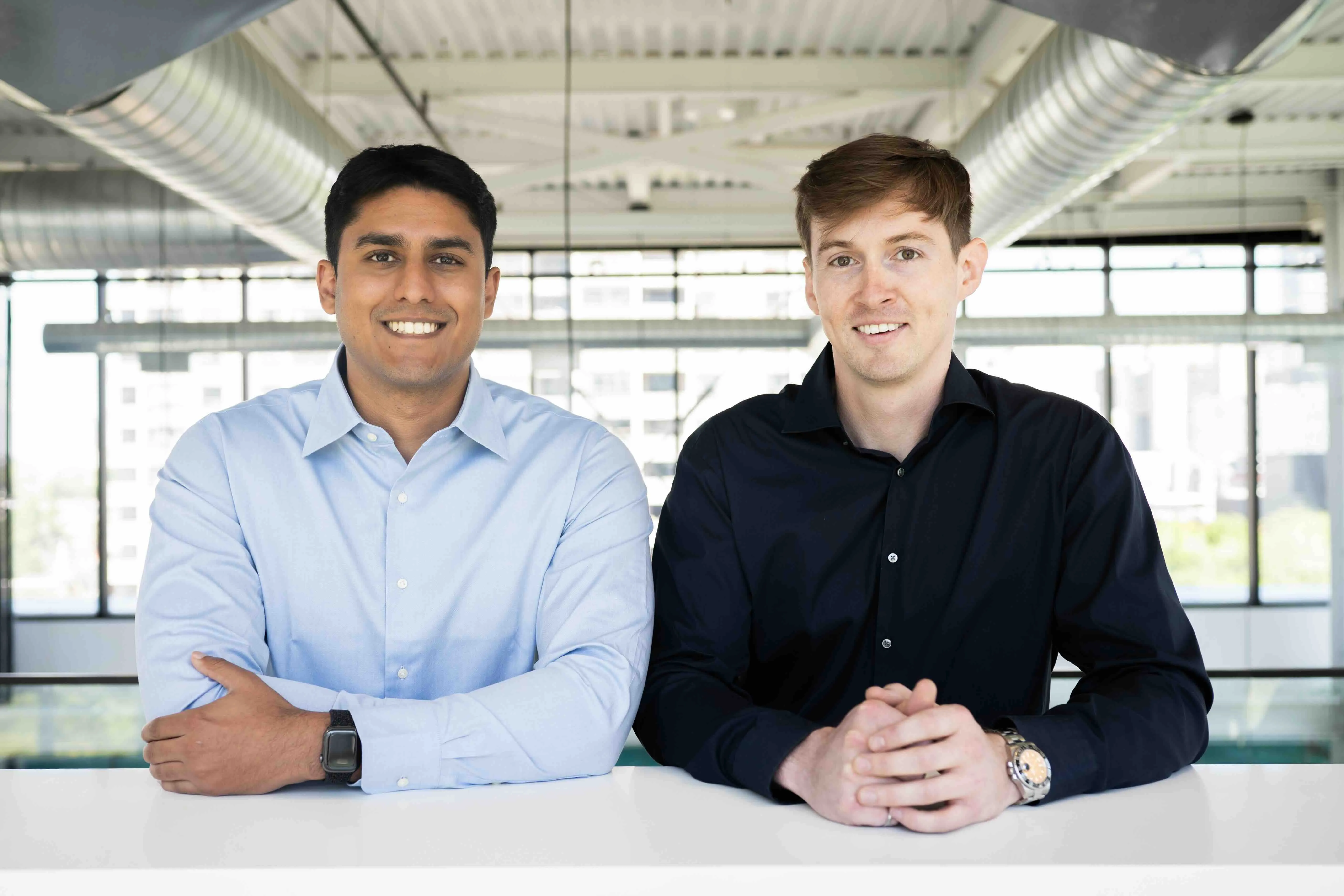 Two men smiling, one in a light blue shirt, the other in a black shirt, sitting at a white table.