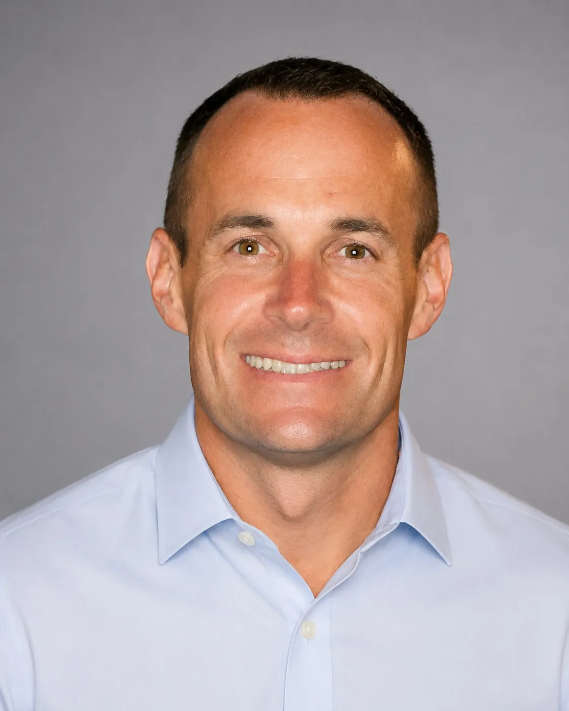 Smiling man with short dark hair wearing a light blue button-up shirt against a gray background.