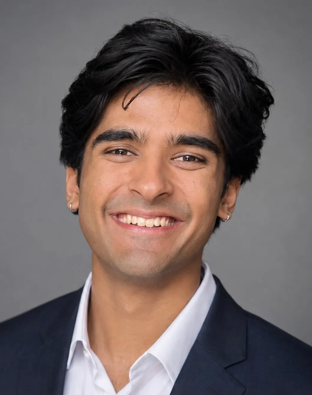 Smiling young man with dark hair, wearing a navy suit jacket and white shirt against a gray background.