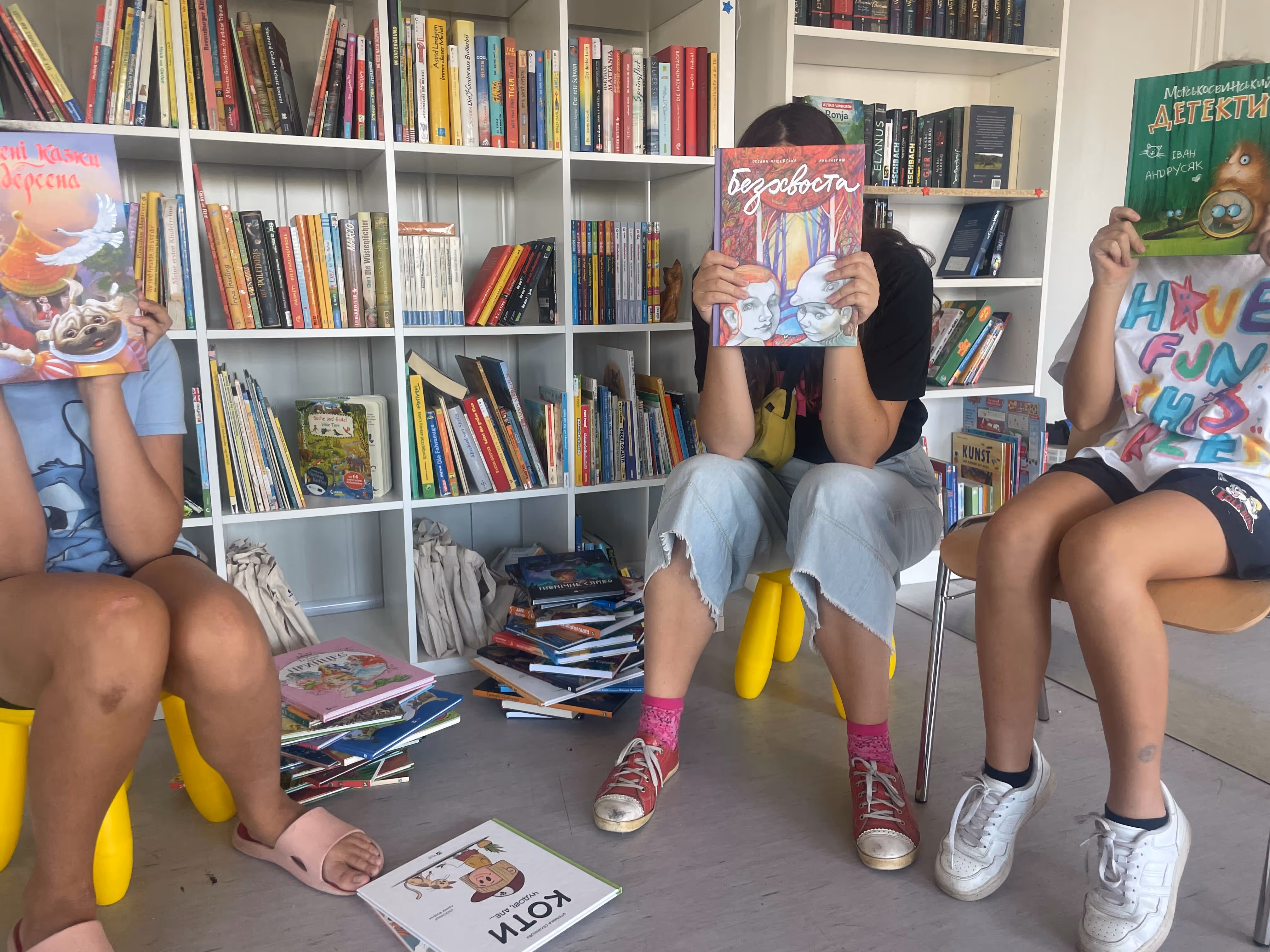 Trois enfants assis dans une bibliothèque, tenant des livres colorés devant leur visage, avec des étagères et des piles de livres derrière eux.