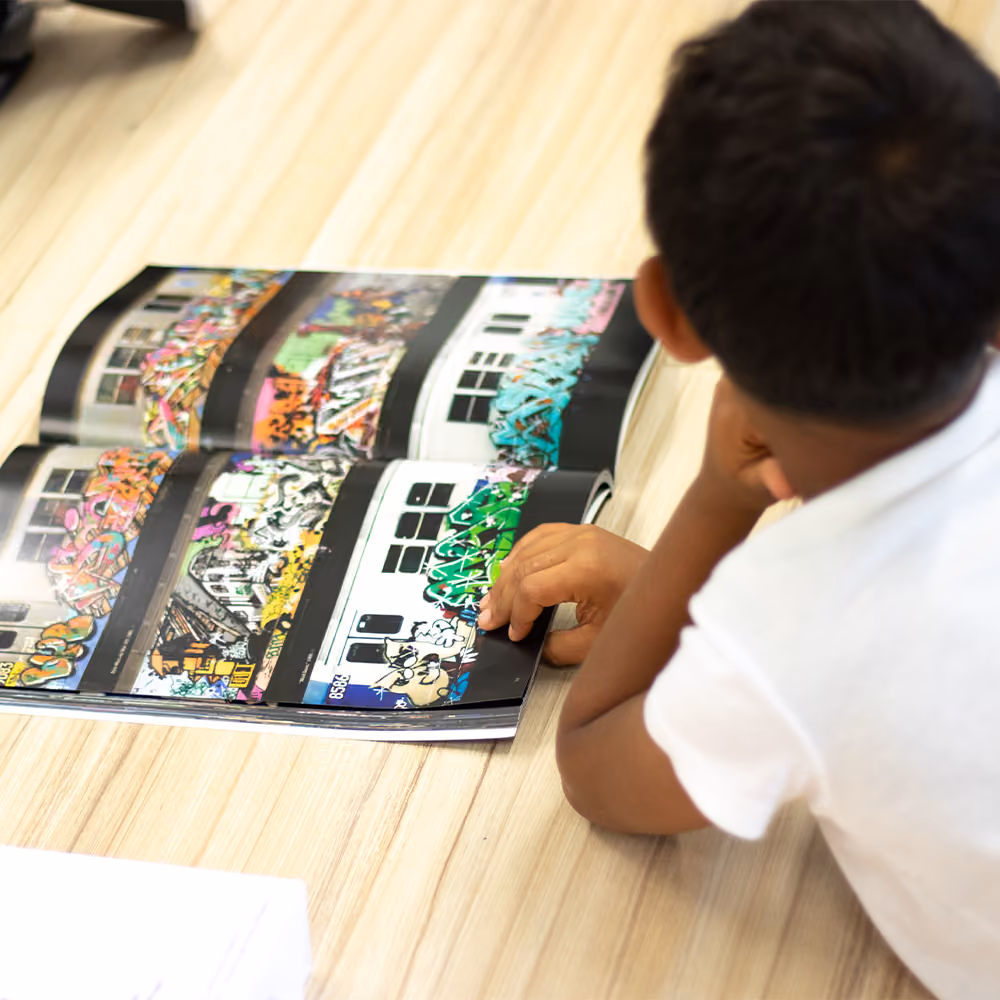 Child lying on a wooden floor looking at a magazine featuring colorful graffiti art on buildings.
